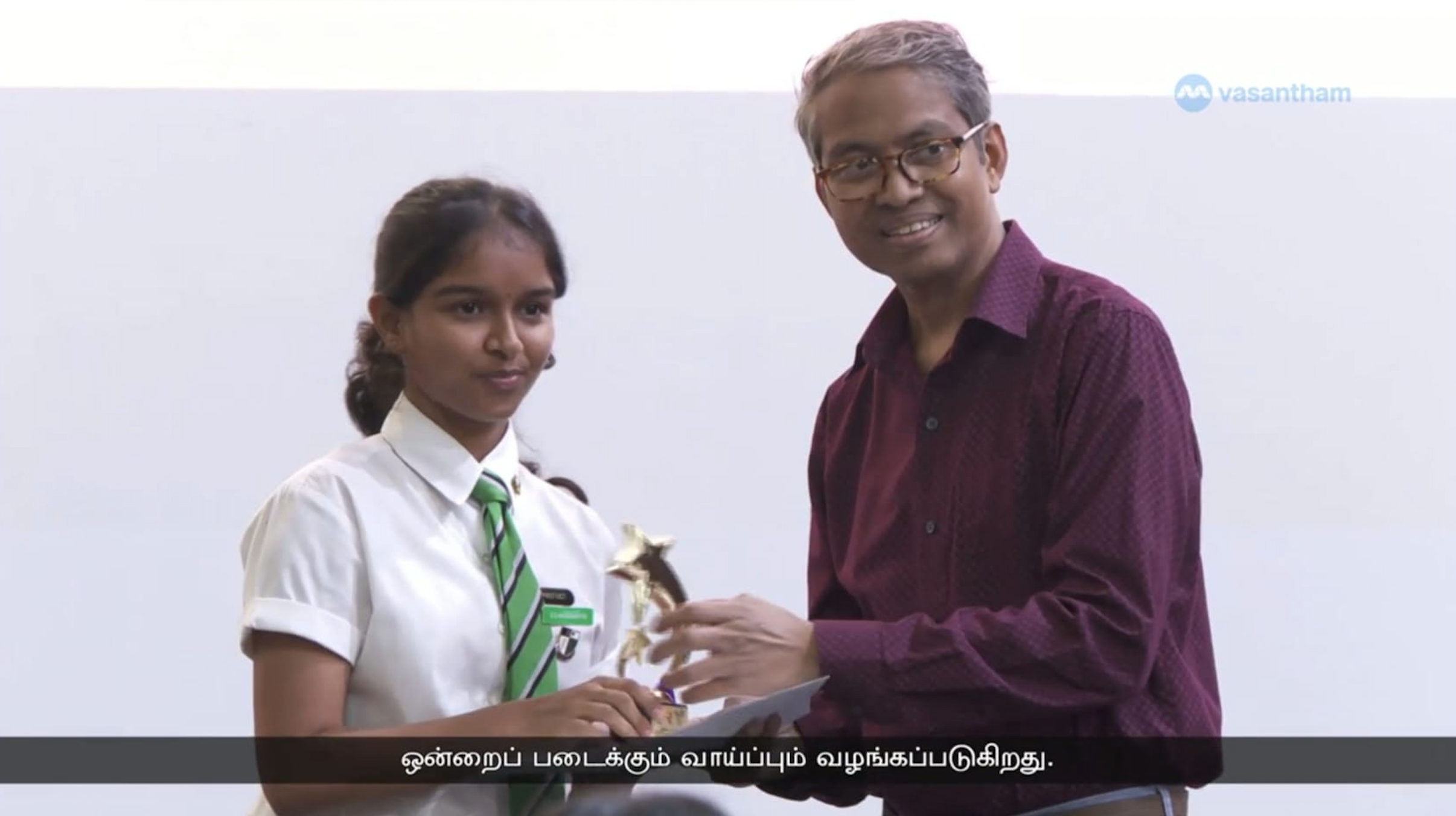 A screen capture of the Vasantham news telecast of a female student being presented a trophy by another man, as they both smile into the camera.